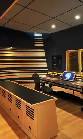 Control room with a mixing console, a studio monitor against a striped timber wall and a window through to the live room
