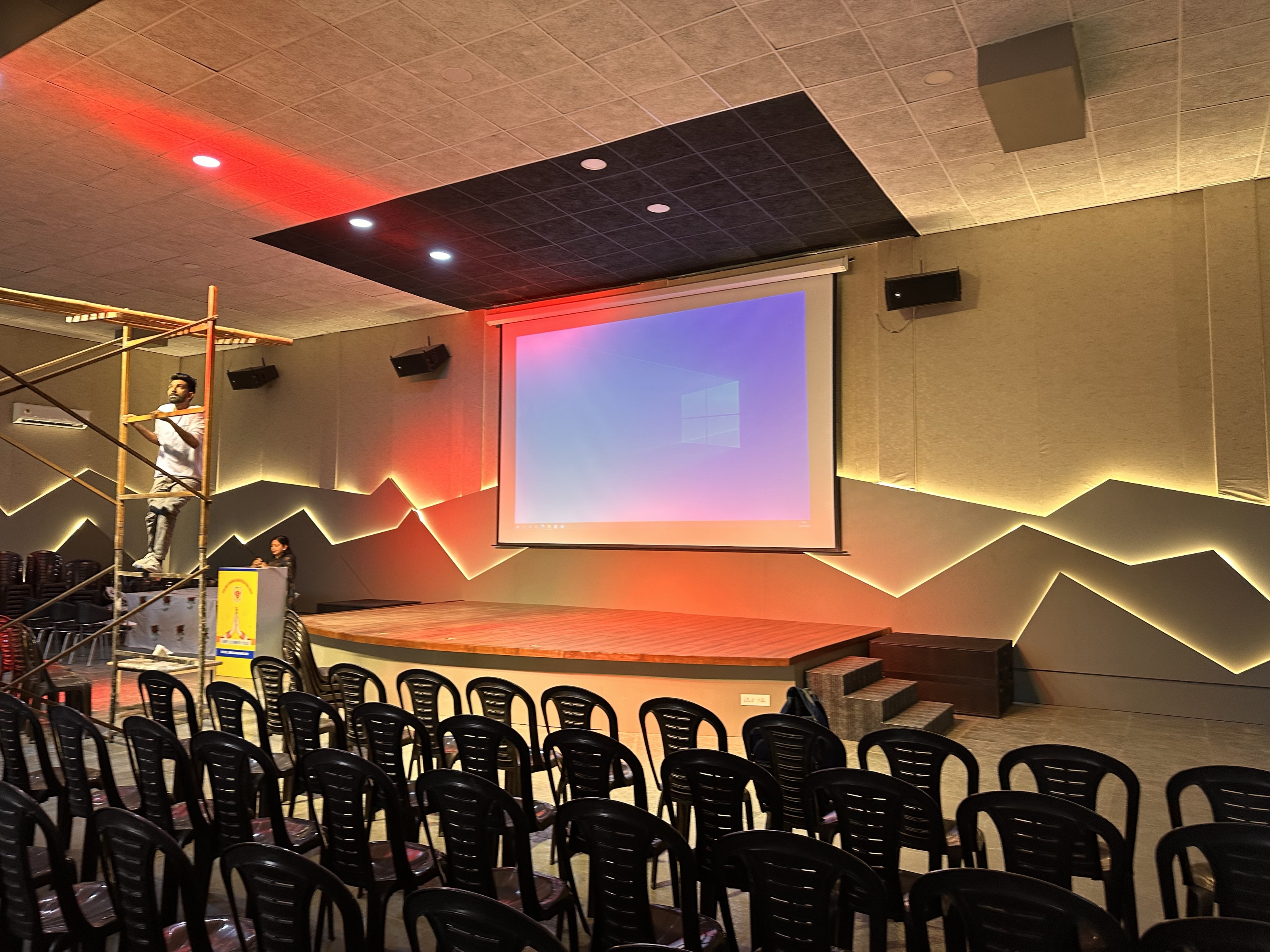 A motorised projection screen lowered above a stage in a school hall, with loudspeakers either side and concealed lighting along the walls.