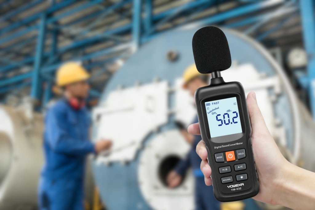 A handheld digital sound level meter reading 50.2 dB, held in front of industrial plant with two workers in hard hats behind.